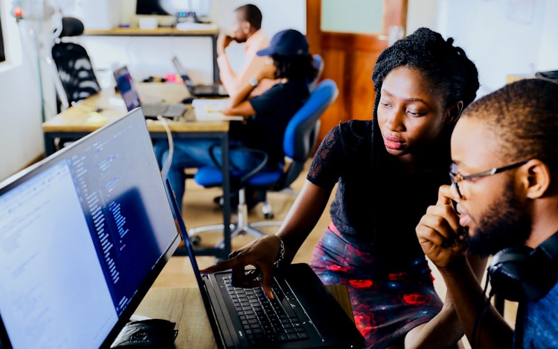 Two colleagues at a desk reviewing code on a monitor in an open-plan office