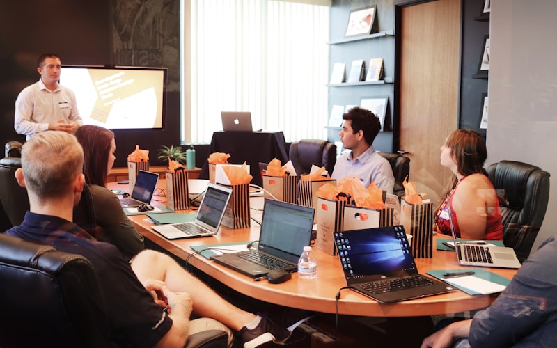 A presenter standing at the front of a boardroom addressing colleagues seated with laptops