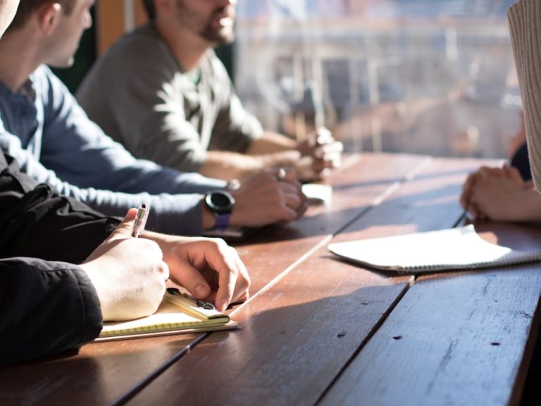 Four people seated around a wooden table, taking notes in notebooks during a working session