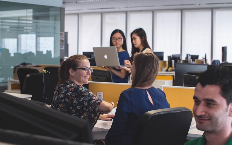 Colleagues talking across desks in an open-plan office, two standing with a laptop