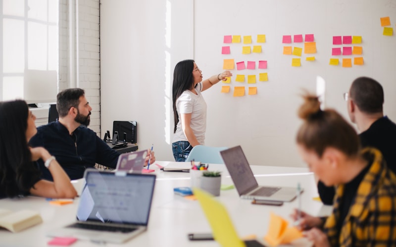 A facilitator pointing at a wall of sticky notes while a seated team works at laptops