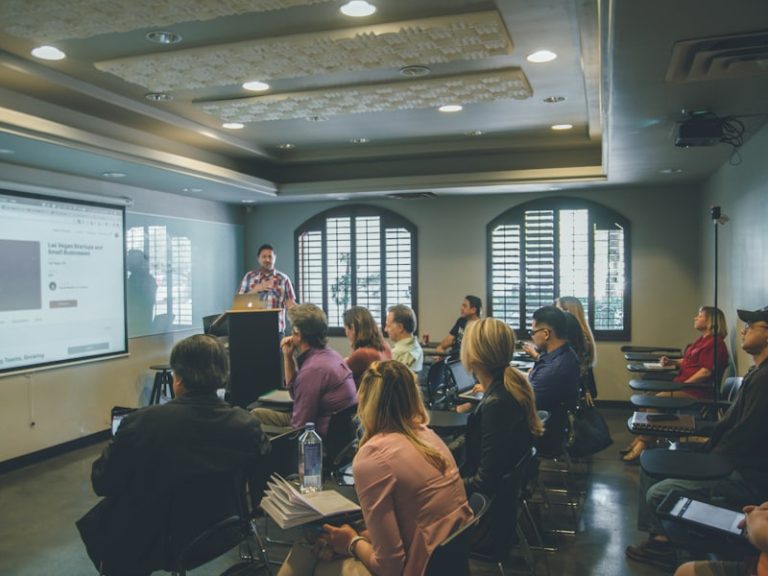 A presenter speaking beside a projection screen to a seated audience in a meeting room